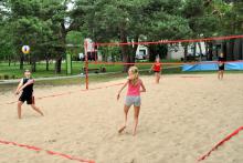 Boisko do gry w piłkę plażową z wyznaczonymi liniami do gry oraz założoną siatką. Dwie pary starszych dzieci gra w siatkówkę. Wokół rosną wysokie drzewa. 