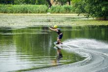 Staw, na środku którego mężczyzna uprawia wakeboarding. Ma na sobie kamizelkę ratunkową, kask na głowie, na nogach przypiętą podłużną deskę, dzięki której może ślizgać się po tafli wody. W rękach trzyma uchwyt połączony z liną, która go ciągnie. 