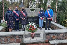 pięć osób stojących obok kamiennego pomnika. Na pomniku ułożona biało-czerwona wiązanka kwiatów.