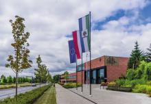 Duży, nowoczesny budynek użyteczności publicznej  ukazany z dużej perspektywy narożnika, ceglane ściany, duże okna i oszklenia, przed budynkiem drzewa oraz horągiew Konstancina-Jeziorny, flagi Polski i Unii Europejskiej na masztach 