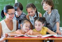 Pięcioro uczniów ogląda z nauczycielką dwie  otwarte książki położone na biurku, w tle zielona tablica szkolna.