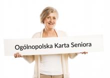 Na zdjęciu uśmiechnięta seniorka, która w rękach trzyma dużą kartę papieru, na której znajduje się napis - Ogólnopolska Karta Seniora. 