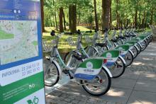 rowery miejskie ustawione na stacji w szeregu. Na pierwszym planie widać fragment tablicy informacyjnej. W tle Park Zdrojowy