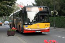 Przód autobusu miejskiego linii 710, jadącego po ulicy, z prawej strony chodnik na którym stoją donice z kwiatami i latarnie