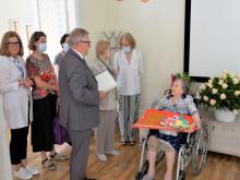 W głównej części mężczyzna, który stoi i trzyma w ręku list gratulacyjny. Obok niego w sali, na wózku inwalidzkim, siedzi starsza kobieta. W tle widać kosze kwiatów oraz inne stojące osoby. 