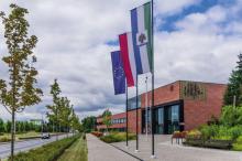 Duży, nowoczesny budynek użyteczności publicznej  ukazany z dużej perspektywy narożnika, ceglane ściany, duże okna i oszklenia, przed budynkiem drzewa oraz horągiew Konstancina-Jeziorny, flagi Polski i Unii Europejskiej na masztach.
