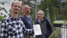 Trzech mężczyzn stoi obok siebie, jeden trzyma w ręku książkę, którą wspólnie napisali. Od lewej: Adam Zyszczyk, Paweł Komosa oraz Witold Rawski. W tle widać wysokie zielone drzewa.