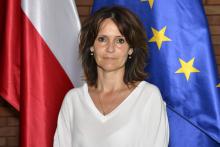 Zdjęcie portretowe kobiety – ciemne proste włosy, ubrana w jasną bluzkę. W tle flaga Polski i Unii Europejskiej.