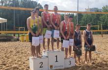 Podium sportowe. Na podestach z numerem 1,2,3 stoją w parach zawodnicy. Na szyi mają medale. W rękach torby z upominkami. W tle widać boisko do siatkówki plażowej. 