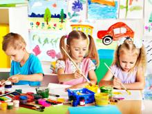 Trójka przedszkolaków siedzi przy stoliku i maluje kolorowymi farbami. W tle galeria z dziecięcymi pracami.