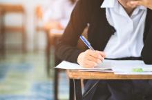 Stolik, przy którym siedzi uczeń zdający egzamin. Przed nim leżą kartki z testem, w prawej ręce trzyma długopis.