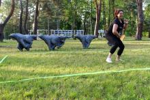 Kobieta w czarnej koszulce i czarnych spodniach podczas gry w crossmintona w parku. W lewej ręce trzyma rakietkę, prawą – ma ugiętą i skierowaną do góry. W tle cztery rzeźby biegnących wilków.