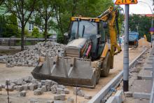 Budowa ulicy z kostki brukowej. W głównej części zdjęcia koparka z opuszczoną łyżką. Do około niej leżą kostki betonowe.  