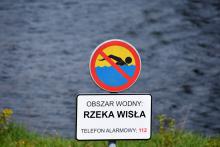Znak zakazujący kąpieli z tabliczką informacyjną, na której jest tekst: obszar wodny – rzeka Wisła, telefon alarmowy 112. W tle nieostre fale płynącej rzeki.  