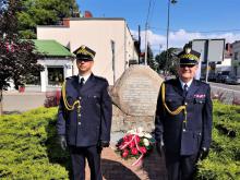 Dwóch strażników miejskich, ubranych w stroje galowe, stoi na baczność obok pomnika upamiętniającego bohaterów Bitwy Warszawskiej z 1920 roku. Pod pomnikiem zostały złożone biało czerwone kwiaty.