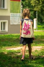 Kilkuletnia dziewczynka idzie po trawie. Na plecach ma jasny worek z napisem kultura, edukacja i zabawa. 