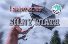 Na górze czerwony napis meteo alert poprzedzony wykrzyknikiem. Obok niego logo Instytutu Meteorologii i Gospodarki Wodnej - niebieski okrąg z białymi literami IMGW, poniżej szary napis silny wiatr. W tle postać z rozwianymi włosami próbująca utrzymać wygięty w górę parasol.  