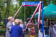 Trzy osoby dokonujące odsłonięcia umieszczonej na pionowym slupie poziomej tabliczki z nazwą „Skwer im. prof. Mariana Weissa”, kobieta w niebieskiej sukience widoczna tyłem, po jej lewej stronie młody mężczyzna w garniturze, na wprost starszy mężczyzna w garniturze, wszyscy pociągają za końcówki biało-czerwonej szarfy, obok stoi dziecko pod parasolem, w tle duży namiot z zielonym dachem, pod którym stoją inne osoby.