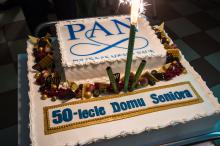 Na stoliku stoi duży biały tort z napisem Polska Akademia Nauk oraz 50-lecie Domu Seniora. Dookoła są czekoladowe ozdoby.