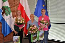 Trzy osoby stoją obok siebie w rzędzie – młoda dziewczyna z długimi blond włosami, kobieta w średnim wieku oraz młoda kobieta. Każda w rękach trzyma bukiet kolorowych kwiatów i torbę prezentową ze zdjęciem Urzędu Miasta i napisem Konstancin-Jeziorna.