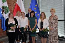  Sala konferencyjna. Siedem osób stoi obok siebie w rzędzie – sześć kobiet i jeden mężczyzna. Cztery kobiety w rękach trzymają dyplomy i białe róże. W tle trzy flagi: Konstancina-Jeziorny, Polski i Unii Europejskiej.