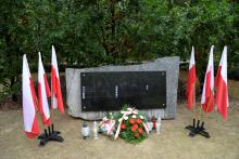Park. Pomnik ofiar katyńskich. Przy nim stoją znicze oraz leżą: biało-czerwone kwiaty i wiązanka z białymi kwiatami. Po obu stronach pomnika stoją na stojaku trzy biało-czerwone flagi. W tle zielone drzewa.