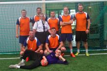 Zdjęcie grupowe drużyny piłkarskiej. 8 zawodników w pomarańczowych koszulkach stoi w bramce. Między nimi jest jedna kobieta ubrana w mundur galowy Ochotniczej Straży Pożarnej. 
