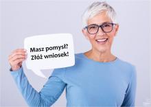 Na zdjęciu uśmiechnięta kobieta – seniorka (krótkie siwe włosy, czarne okulary, ubrana jest w niebieską bluzkę), kobieta trzyma tabliczkę, z napisem: w pierwszym wierszu: Masz pomysł? w drugim wierszu: Złóż wniosek!