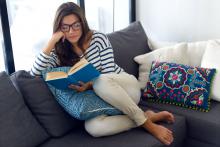 Młoda kobieta siedzi na kanapie, w ręku trzyma i czyta książkę, na nosie ma okulary. Na kanapie leżą poduszki.