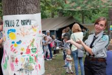 Kobieta w mundurze harcerskim gestem rąk zaprasza do obozu harcerskiego, w tle kilkadziesiąt osób oraz zielony namiot. Na drzewie wisi kolorowy obrazek z napisem: Maluj z nami.