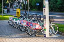 Stacja wypożyczalni rowerów miejskich z kilkunastoma rowerami. W tle ulica oraz jadąca po chodniku rowerzystka.