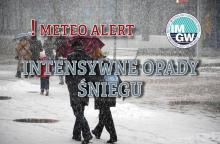 Na górze czerwony napis meteo alert poprzedzony wykrzyknikiem. Obok niego logo Instytutu Meteorologii i Gospodarki Wodnej - niebieski okrąg z białymi literami IMGW, poniżej szary napis intensywne opady śniegu. W tle widać ludzi idących chodnikiem. Pada na nich śnieg. 