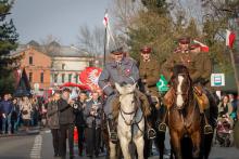 Przemarsz ulicą. Na pierwszym planie trzech mężczyzn w mundurach jadą na koniach, za nimi maszeruje orkiestra dęta oraz ludzie. 