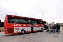 Na chodniku stoi duży czerwony autobus z białym napisem z boku Mobilny Punkt Poboru Krwi. Przed autobusem stoi sześć osób. 