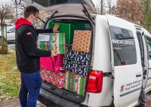 Samochód dostawczy, który ma otwarty bagażnik. Znajdują się w nim duże pudła – prezenty świąteczne. Obok samochodu stoi mężczyzn, który pakuje do niego kolejną paczkę.