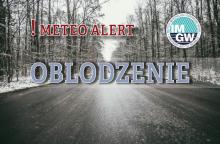 Na górze czerwony napis meteo alert poprzedzony wykrzyknikiem. Obok niego logo Instytutu Meteorologii i Gospodarki Wodnej - niebieski okrąg z białymi literami IMGW, poniżej szary napis oblodzenie. W tle widać samochód jadący ulicą asfaltową przez las.  