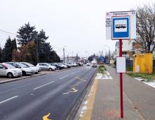 Ulica. Po prawej stronie znajduje się chodnik. Na nim stoi wysoki słup z tabliczką – nazwą przystanku autobusowego i rozkładem jazdy. 