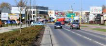 Ulica dwukierunkowa w Konstancinie-Jeziornie. Po ulicy jadą samochody, po chodnikach idą piesi. W dali widać budynki oraz duże banery reklamowe. 