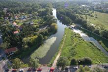 Zdjęcie miasta z lotu ptaka. W dolnej części widać ulicę i jadące po niej samochody. Obok ulicy płynie rzeka. W dali widoczne są zabudowania jednorodzinne. 