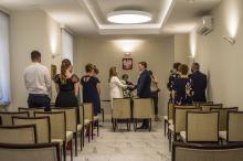 Sala ślubów cywilnych. Na środku naprzeciwko siebie stoi para młoda – trzymają się za ręce, składają przysięgę. Po bokach sali stoją goście – około 10 osób. 