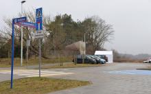 Parking z kostki brukowej. Poi lewej stronie widoczny drogowskaz z nazwami ulic. W tle kilka zaparkowanych samochodów. Na końcu parkingu stoi biały duży namiot. 