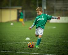 Dziecko – chłopak, ubrany w strój sportowy biegnie po boisko kopiąc piłkę. 