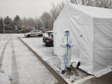 Parking samochodowy z kostki brukowej. Po prawej stronie widoczny biały duży namiot, przed którym stoi osoba w białym kombinezonie i maseczce, z tyłu stoją dwa samochody osobowe.