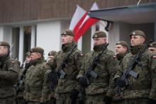 Grupa kilkunastu żołnierzy w umundurowaniu i z bronią automatyczną przewieszoną na piersiach, na głowach berety, za nimi widoczny fragment budynku z umocowanymi dwiema flagami biało-czerwonymi.