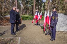Trzech mężczyzn ubranych na czarno stoi przed pomnikiem katyńskim – dużym kamieniem, na którym wisi granitowa tablica. Pod tablicą leżą kwiaty i palą się znicze. 