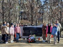 Grupka dzieci oraz dorosłych stoi obok pomnika katyńskiego – dużego kamienia, na którym wisi granitowa tablica. Pod tablicą leżą kwiaty i palą się znicze.