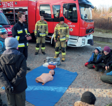 Trzech stojących strażaków na tle wozu strażackiego. Dookoła grupa młodzieży, na ziemi leży niebieska mata, a na niej manekin do ćwiczeń z pierwszej pomocy.