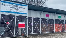 Na zdjęciu metalowa podłużna wiata. Pod nią stoi sześć dużych kontenerów budowlanych. Na wiacie powieszony jest duży baner z napisem: Gminny Punkt Selektywnej Zbiórki Odpadów Komunalnych.