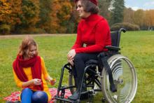 Polana, w tle las jesienią. Na wózku inwalidzkim siedzi kobieta z niepełnosprawnością (uśmiecha się). Towarzyszy jej druga kobieta, która siedzi na kocu rozłożonym na ziemi.