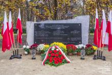 Pośrodku czarna tablica granitowa z tekstem umocowana na prostokątnym kamieniu, po lewej i prawej stronie stoją flagi narodowe, przed tablicą leżą kwiaty i wiązanki oraz palą się znicze